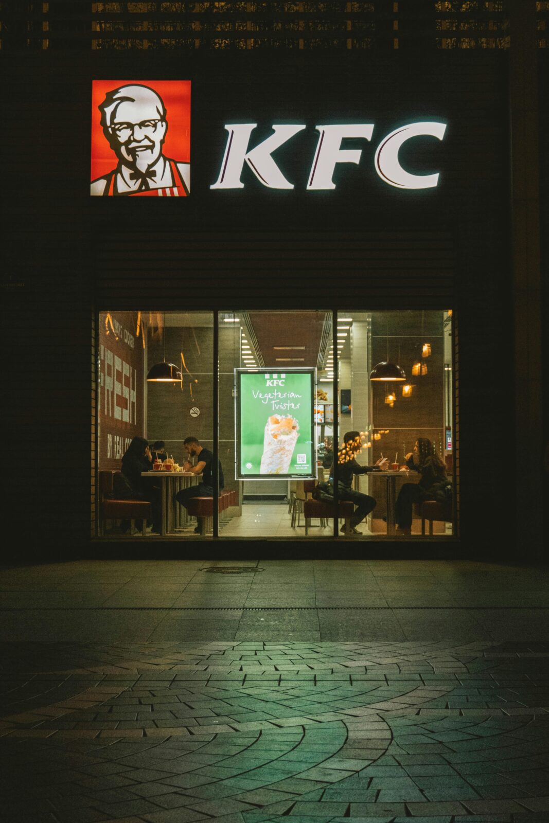 Nighttime view of a KFC storefront with diners visible through the glass facade.