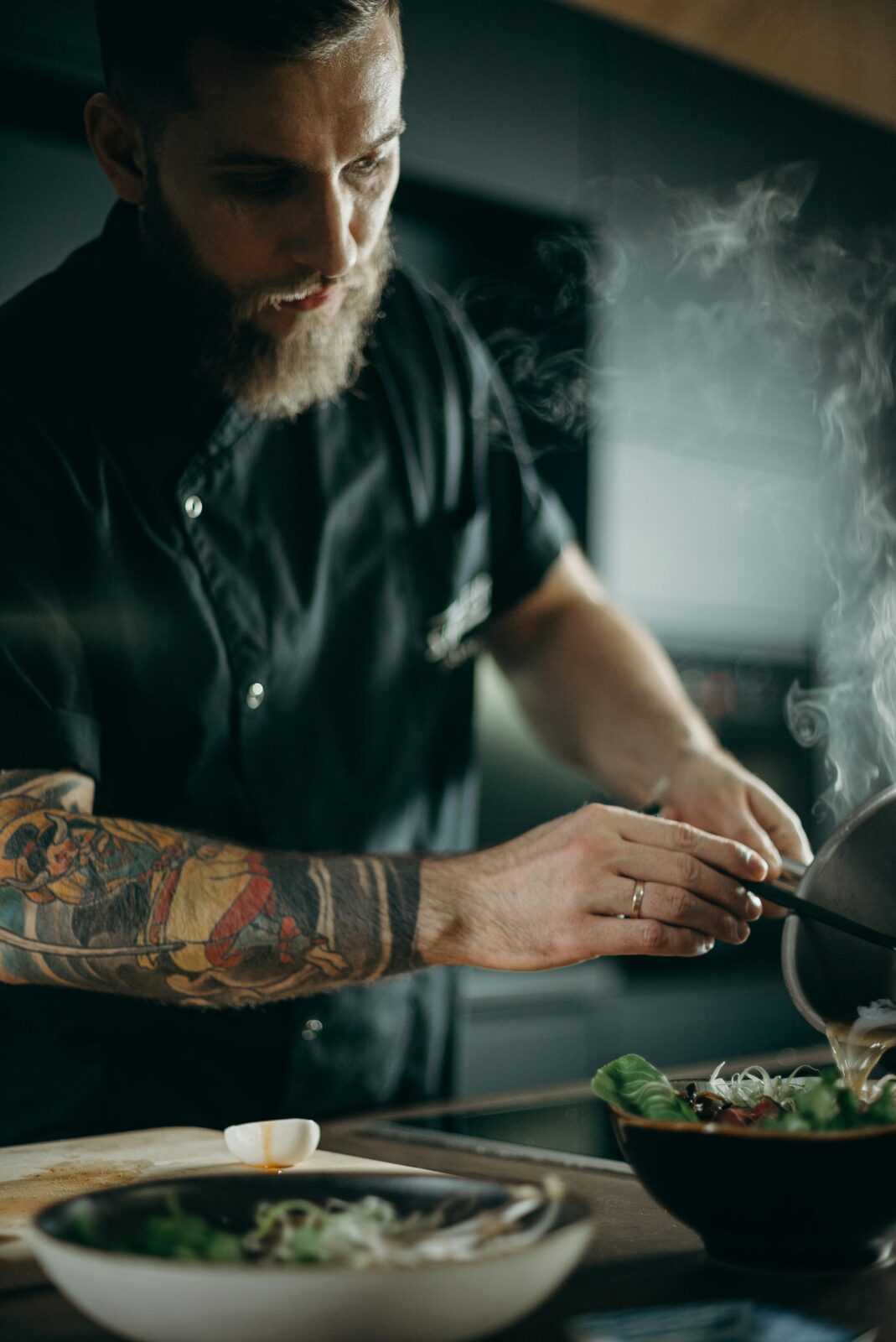 Chef with tattoos pouring hot soup into a bowl with steam rising.