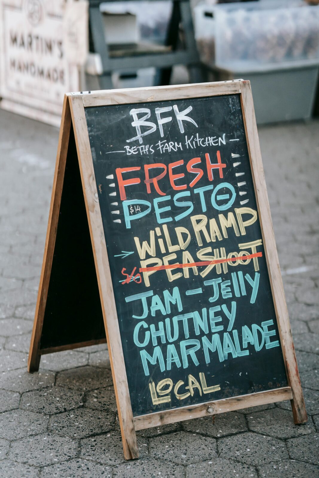 Colorful chalkboard sign at outdoor market listing fresh farm products like pesto, wild ramp, and more.